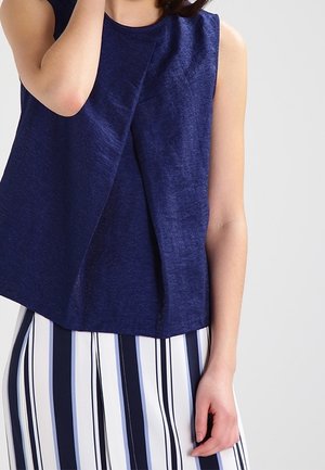 Bezrękawnik w ciemnym granacie z plisą z przodu, noszony z białymi spodniami z pionowymi granatowo-czarnymi paskami.