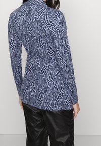 Dlouhá rukávová blůza v námořnické modři s tečkovaným vzorem. Tkanina má hladkou texturu. Střih je přiléhavý v pase, s vysokým límcem.