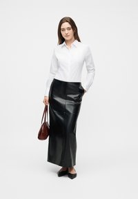 Γυναίκα με λευκό πουκάμισο με κουμπιά και μακριά μαύρη δερμάτινη φούστα, κρατώντας καφέ τσάντα, στέκεται μπροστά σε απλό λευκό φόντο.