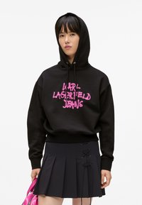 Persona giovane che indossa un hoodie nero con il testo rosa "Karl Lagerfeld Jeans", una gonna nera plissettata con dettagli in lacci, e tiene una borsa rosa.