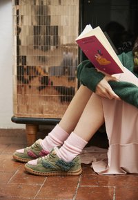 Grüne Wildledersneaker mit strukturierten rosa und cremefarbenen Akzenten, kombiniert mit hellrosa gerippten Socken, die auf einem Fliesenboden ruhen. Ein Buch liegt in der Nähe.