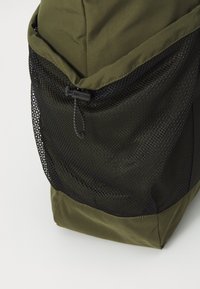 Sac à dos vert avec une poche en maille noire sur le côté, présentant une texture de tissu lisse et une fermeture à cordon pour un rangement sécurisé.
