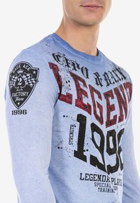 Himmelblaues Langarmshirt mit auffälligen schwarzen und roten Grafiken, darunter "LEGEND" und "1996", sowie einem Aufnäher auf dem linken Ärmels.