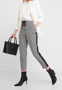 Černobílé kalhoty s potiskem houndstooth a bočními černými pruhy, doplněné bílou blůzou a elegantní černou kabelkou. Černé kožené boty na podpatku.