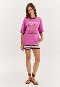 Růžové oversized tričko s grafickým potiskem, spárované s pruhovanými pletenými šortkami v červené a bílé. Hnědé sandály s omotanými pásky kolem kotníků.