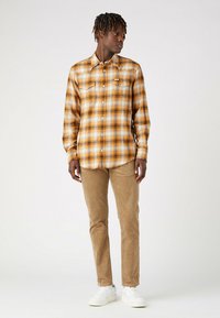 Camicia a maniche lunghe in flanella a quadri gialli e arancioni, abbinata a pantaloni in corduroy beige e sneakers bianche, creando un look casual.