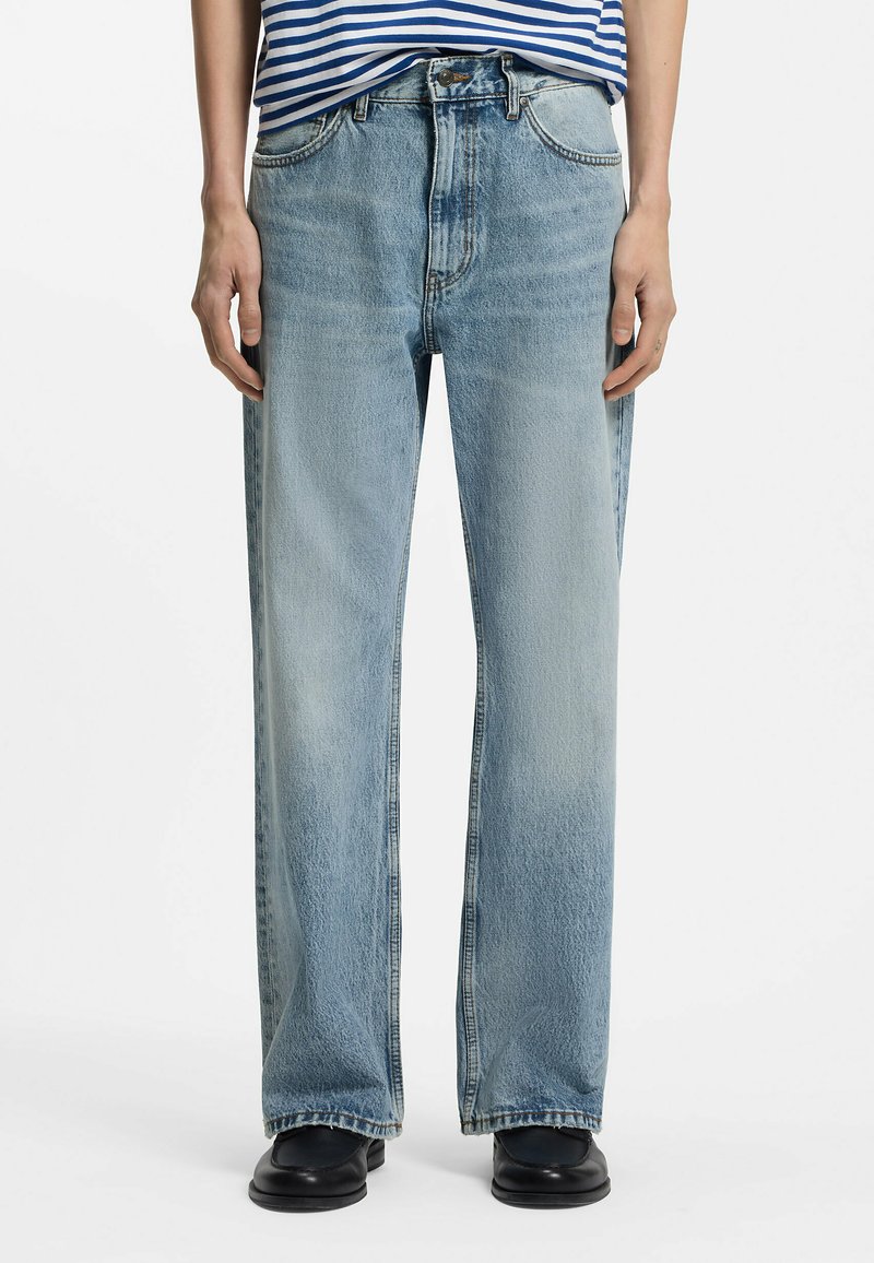 Personne portant un jean bleu clair à jambes droites, des chaussures noires et une chemise rayée bleu et blanc, debout, les bras détendus le long du corps.