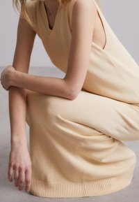 Robe beige sans manches, longueur genou, avec un col en V, une encolure côtelée et un ourlet. Texture douce avec une silhouette ajustée, montrée sur une figure accroupie.