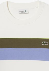 Camiseta de algodón blanca con rayas horizontales en verde olivo, azul claro y un acento en verde neón, con un pequeño logo bordado.
