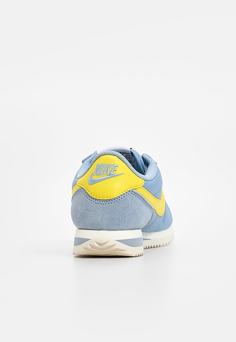Baskets Nike avec une tige en suede bleu clair, des accents jaunes au niveau du talon et du swoosh, et une semelle en caoutchouc blanc texturé.