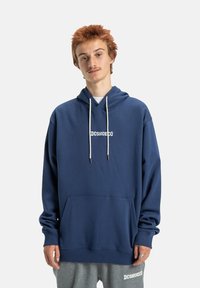 Námornícky modré mikina s kangaroo vreckom, bielymi šnúrkami a potlačou "DCSHOECO". Vyrobená z mäkkej tkaniny s voľným strihom.