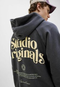Gray hoodie with large cream lettering on the back, featuring "Studio Originals" and additional text. Soft fabric with a relaxed fit.