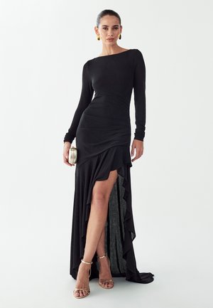 Žena v čiernych šatách s dlhým rukávom a vysokým rozparkom, držiaca striebornú kabelku, s pozlátenými náušnicami a zlatými sandálmi na vysokom opätku, na bielom pozadí.