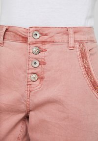 Rosa denimbyxor med hög midja, försedda med fyra dekorativa knappar, avancerad sömnad och en texturerad sidopanel.