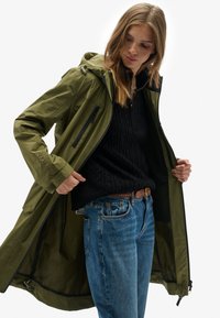 Chaqueta impermeable verde oliva con capucha, bolsillos con cremallera y puños ajustables; combinación con un suéter negro texturizado y jeans azules.