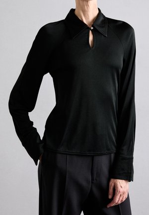 Personne portant un haut noir à manches longues avec une fente étroite et un bouton au col, assorti d'un pantalon noir ample.