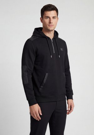 Young man wearing a black hoodie with front pocket and zippered collar, standing against a plain light gray background.