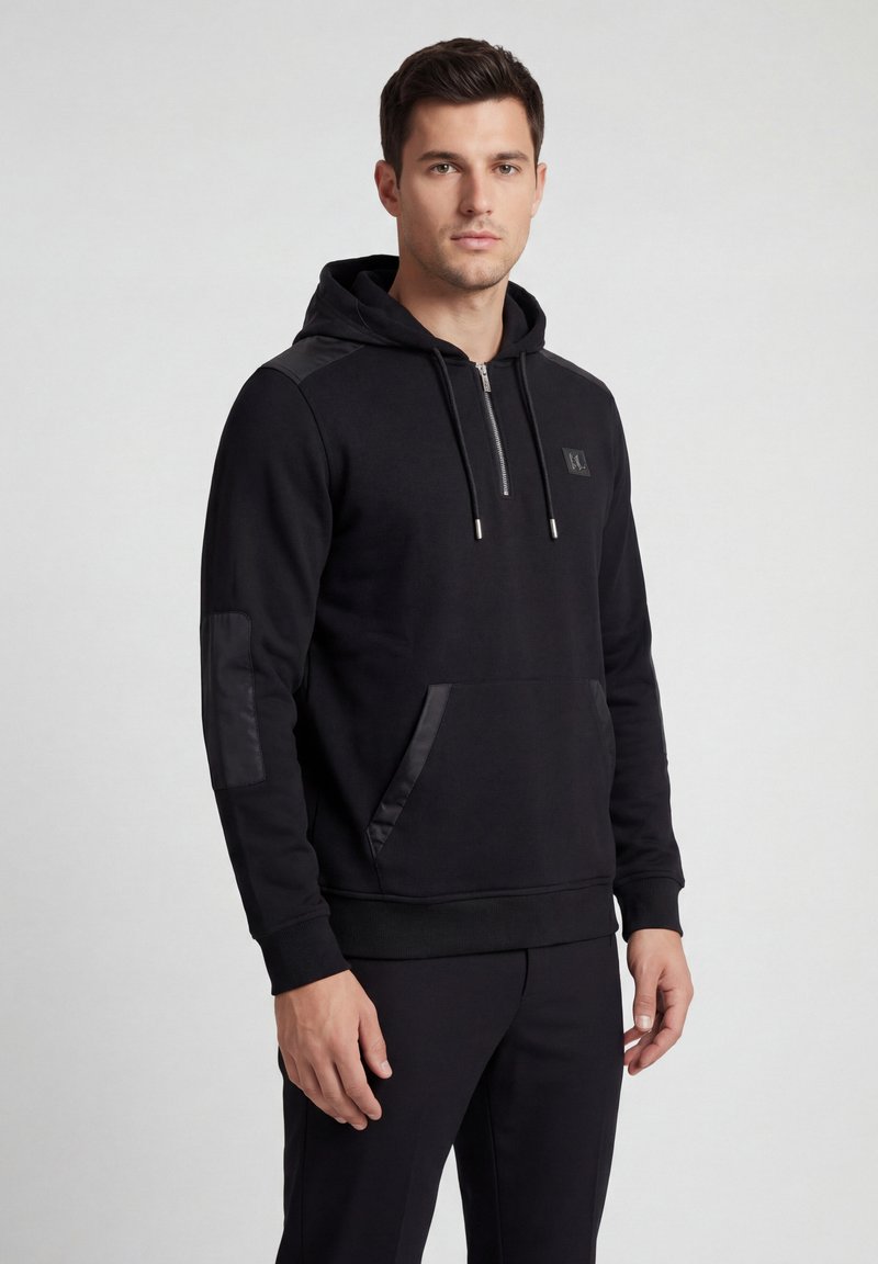 Jeune homme portant un sweat à capuche noir avec poche avant et col zippé, debout devant un fond uni gris clair.