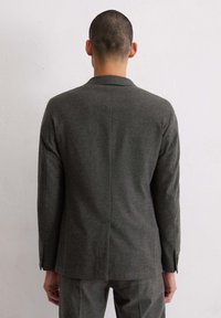 Man met kort haar, gekleed in een donkergrijze op maat gemaakte blazer en bijpassende broek, staande tegenover een effen witte muur.