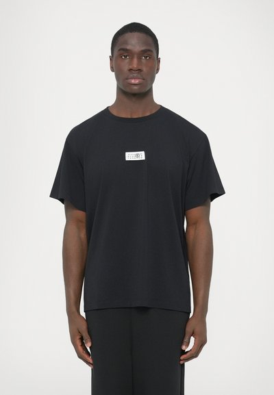 Μαύρο βαμβακερό T-shirt με άνετη γραμμή, κοντά μανίκια και ορθογώνια λευκή ετικέτα στο στήθος με κείμενο και λογότυπο.