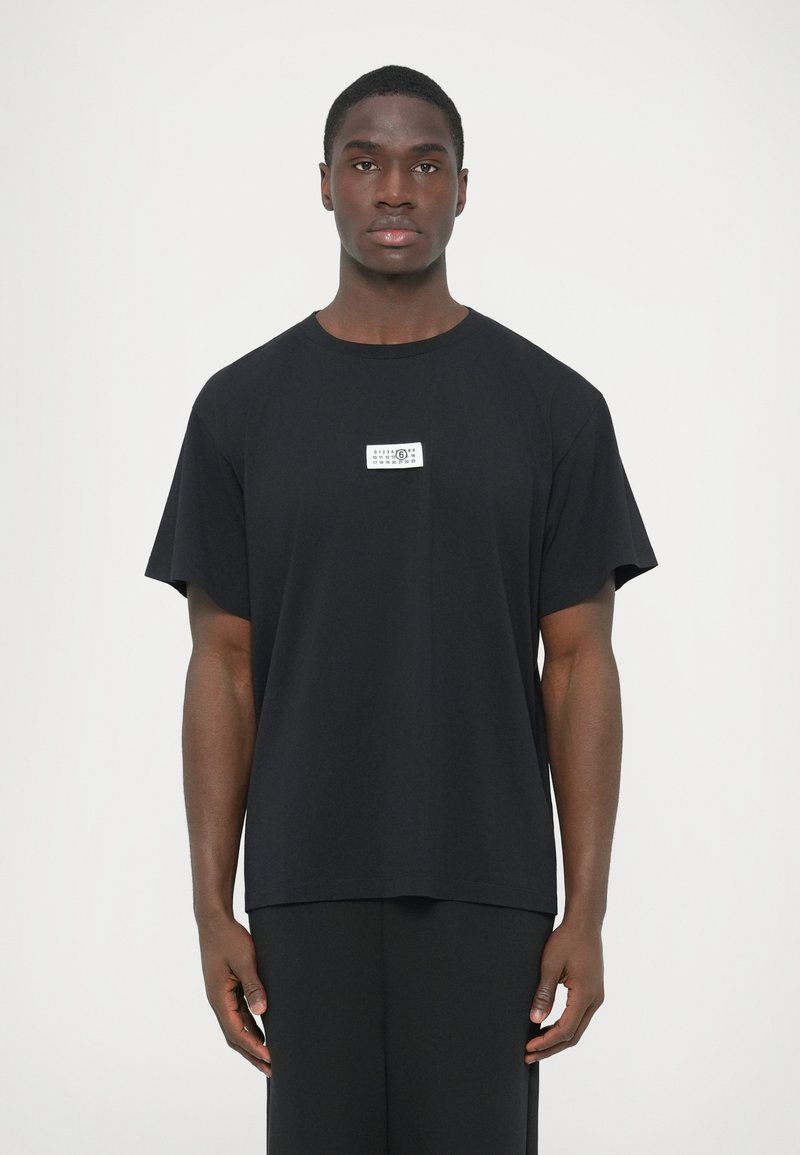 Μαύρο βαμβακερό T-shirt με άνετη γραμμή, κοντά μανίκ�ια και ορθογώνια λευκή ετικέτα στο στήθος με κείμενο και λογότυπο.