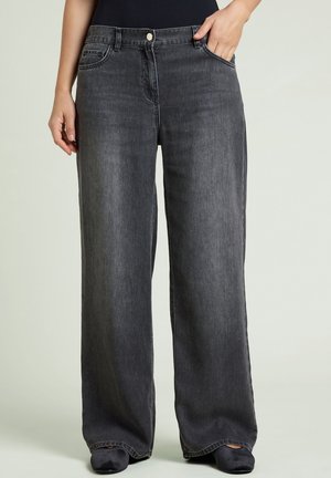 Person som bär löst sittande, mörkgrå denimjeans med framfickor, en knapp och dragkedja, står mot en enkel, ljus bakgrund.