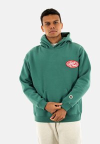 Sudadera verde de gran tamaño, hecha de una tela suave, con un bolsillo frontal, un logo bordado en rojo y puños y dobladillo de canalé.