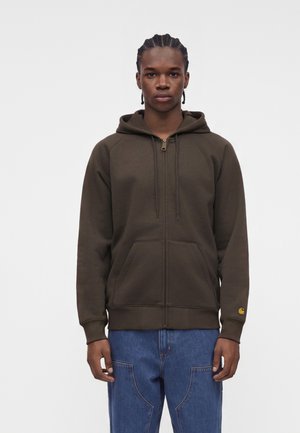 Young man with braided hair wearing a dark brown zip-up hoodie and blue jeans, standing against a plain white background.
