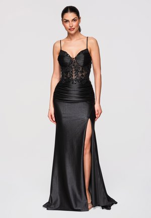 Kvinde iført en gulvlængde sort kjole med tynde stropper, blomsterbroderet gennemsigtig overdel og høj slids, stående mod en ensfarvet baggrund.