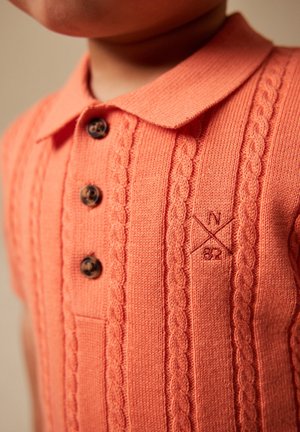Primer plano de un niño que lleva una camisa polo de punto color coral con patrones de cable, tres botones marrones y bordado "N 82" en el pecho.
