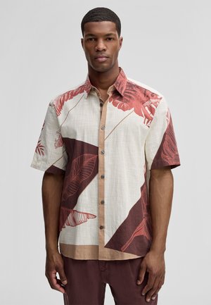 Mann trägt ein kurzärmeliges Hemd mit auffälligen roten und braunen tropischen Blattmustern auf cremefarbenem Stoff.