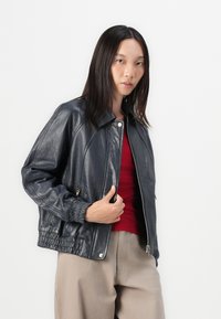 Giacca bomber nera in pelle con chiusura a zip, polsini a costine e texture morbida. Abbinata a un top rosso e pantaloni beige.