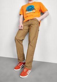 Oranžové bavlněné tričko s modro-černým motivem hor, doplněné světle hnědými chino kalhotami a červenými teniskami s bílými pruhy.