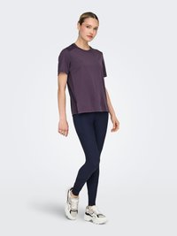 Kortärmelig, paarse sport-T-shirt met gestructureerde schouderpanelen, gecombineerd met marineblauwe legging en wit-zwarte sneakers, staande op een lichte achtergrond.