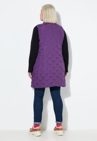 Frau mit kurzen blonden Haaren, die eine lange, lila gesteppte Weste, schwarze Langarmshirts, Bluejeans und weiße Sneakers mit pinken Akzenten trägt, mit dem Rücken zur Kamera.