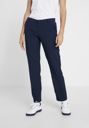 Kobieta ubrana w granatowe spodnie o prostych nogawkach, białe sneakersy z czarnymi podeszwami i biały top bez rękawów, stojąca na jednolitym tle.