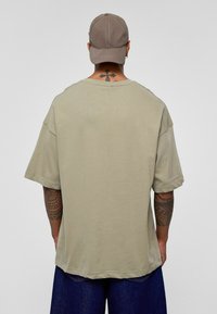 Overmaatse olijfgroene katoenen t-shirt met een ronde hals en korte mouwen, gedragen met blauwe denim shorts, gezien van achteren.