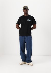 Zwarte katoenen t-shirt met een kleurrijk "Santa Cruz" logo op de borst, gecombineerd met losse blauwe spijkerbroek en beige sneakers.
