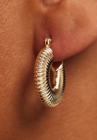 Gouden oorring met geribbelde textuur, cirkelvormig en voorzien van een veilige sluiting, hangend aan een oorlel tegen een huidkleurige achtergrond.