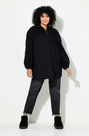 Camisa negra de gran tamaño con botones y mangas largas abullonadas, combinada con jeans oscuros y botas negras hasta el tobillo. Textura de tela suave.