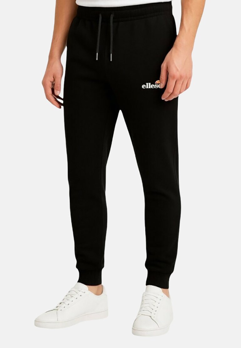 Pantalon de jogging noir avec taille à cordon, arborant un logo blanc sur la cuisse gauche et des poignets fuselés. Porté avec des baskets blanches.