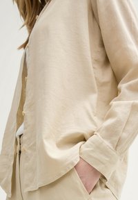 Camicia a maniche lunghe in lino beige con vestibilità comoda, caratterizzata da un davanti con bottoni, una leggera trama e un orlo arrotondato con maniche a sbuffo.