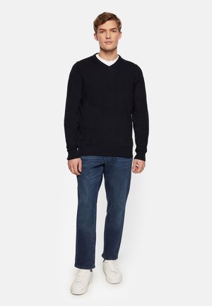 Donker marineblauwe v-hals trui gemaakt van gebreid materiaal, gecombineerd met blauwe jeans en witte sneakers. De trui heeft een geribbelde rand en manchetten.
