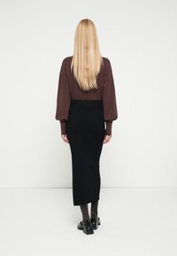 Brązowy sweter z bufkami na rękawach, zestawiony z czarną, prążkowaną spódnicą midi. Modelka nosi czarne buty na obcasie. Gładkie białe tło.