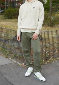 Osoba v béžové mikině Nike, olivově zelených teplácích Nike a bílých teniskách stojící na chodníku poblíž černého kovového plotu a stromů.