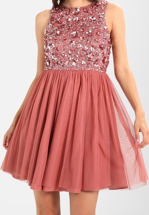Robe rose sans manches avec un corsage fleuri orné de perles et une jupe en tulle à couches jusqu'aux genoux, portée par une personne debout.