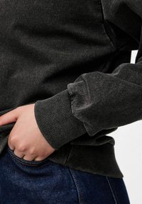 Ranka poilsio tamsiai mėlynuose džinsuose, vilkint juodą ilgomis rankovėmis megztinį su raukiniais ant rankovių ir apatinės dalies.