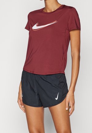 Persoon die een donkerrood Nike T-shirt met korte mouwen en zwarte Nike sportshorts draagt tegen een effen achtergrond.