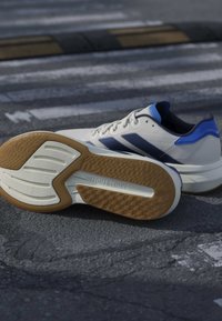 Chaussures de course blanches et bleues sur l'asphalte, une chaussure montrant une semelle texturée marron et blanche avec le texte "LIGHTSTRIKE".