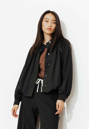 Femme aux longs cheveux bruns portant une veste noire ample à boutons, un haut marron, et un pantalon noir avec un cordon blanc, debout devant un fond blanc.